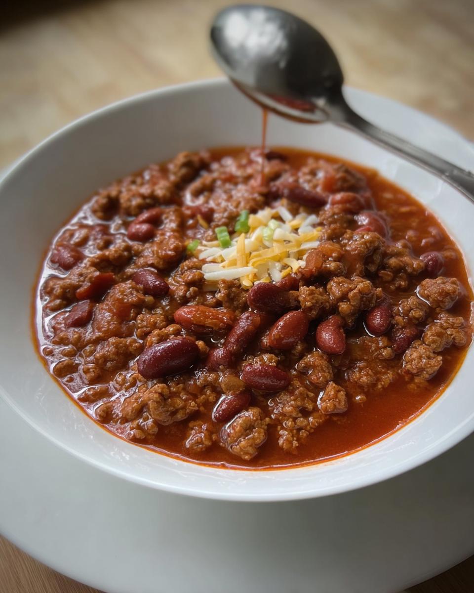 A close-up of a bowl of hearty ground beef chili with kidney beans, topped with shredded cheese and green onions.