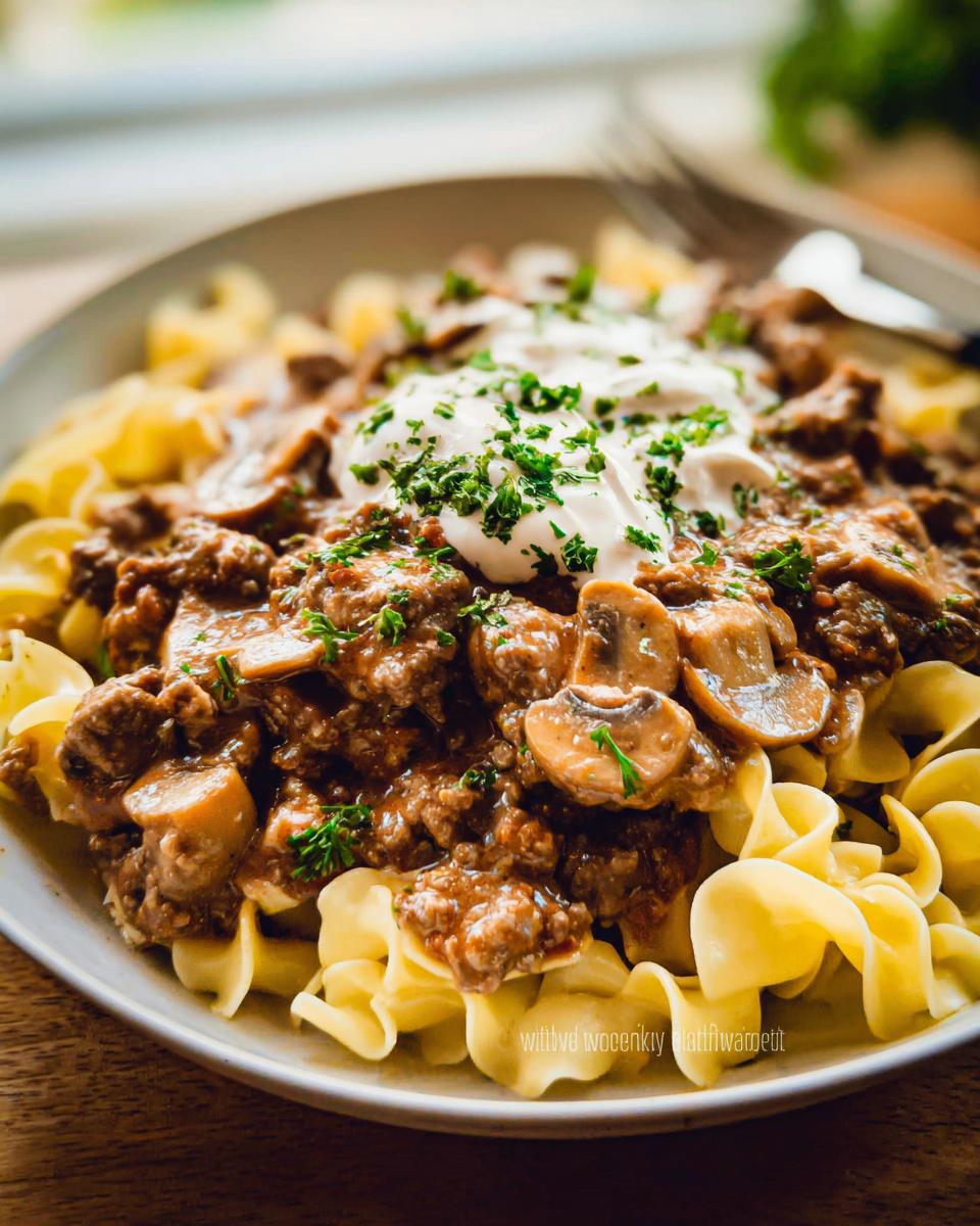 A bowl of creamy beef stroganoff with mushrooms served over egg noodles and topped with sour cream and parsley.