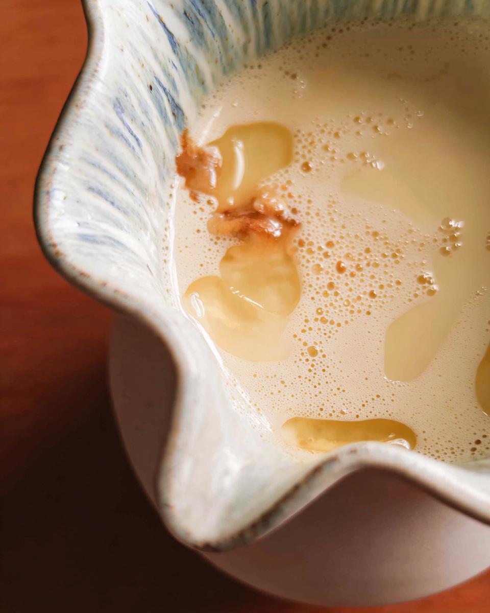 Close-up of a light, frothy Champagne Cream Sauce for Seafood being prepared in a rustic ceramic pitcher.