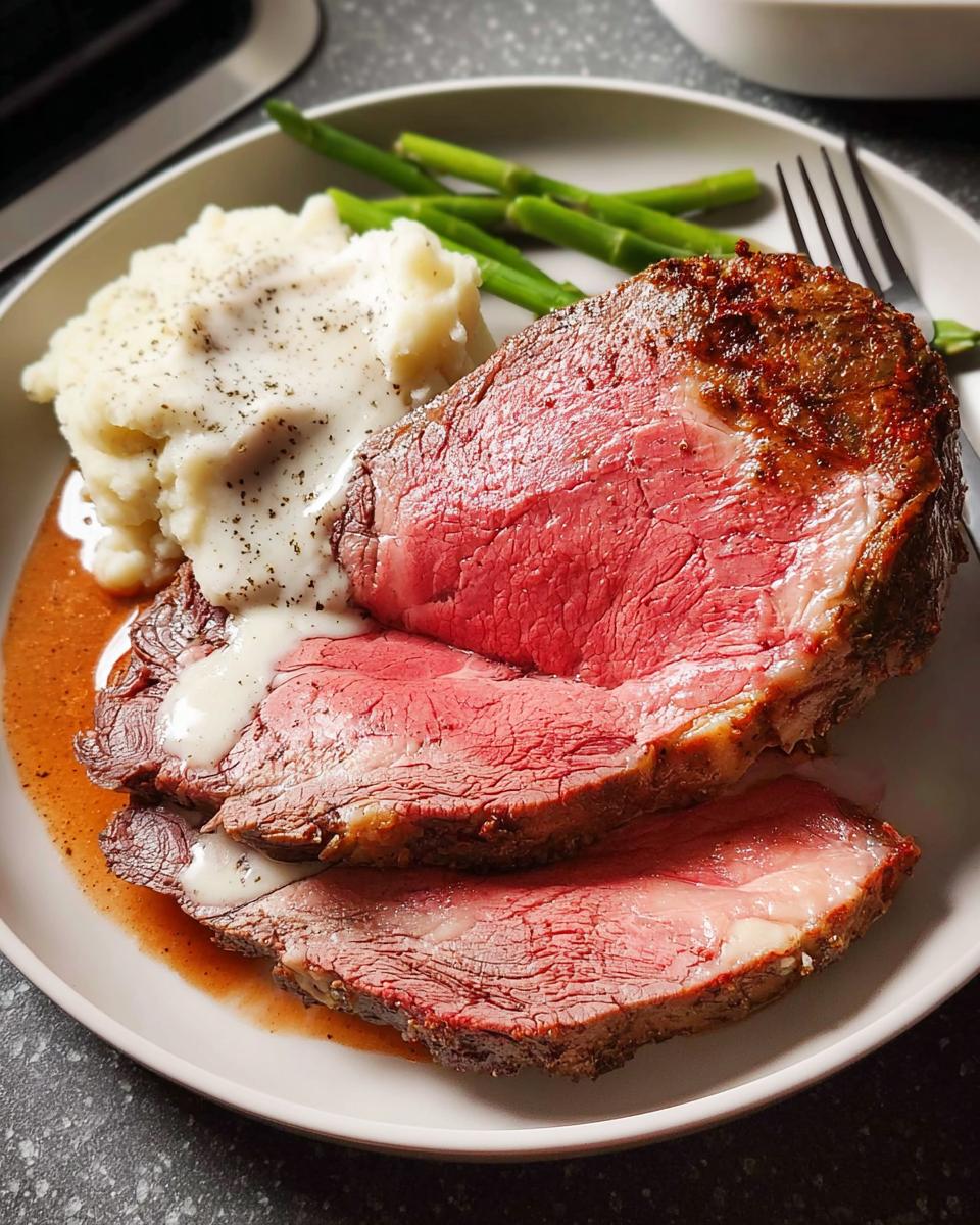 Two thick slices of perfectly cooked Easy Prime Rib roast, medium-rare, served with mashed potatoes and asparagus.