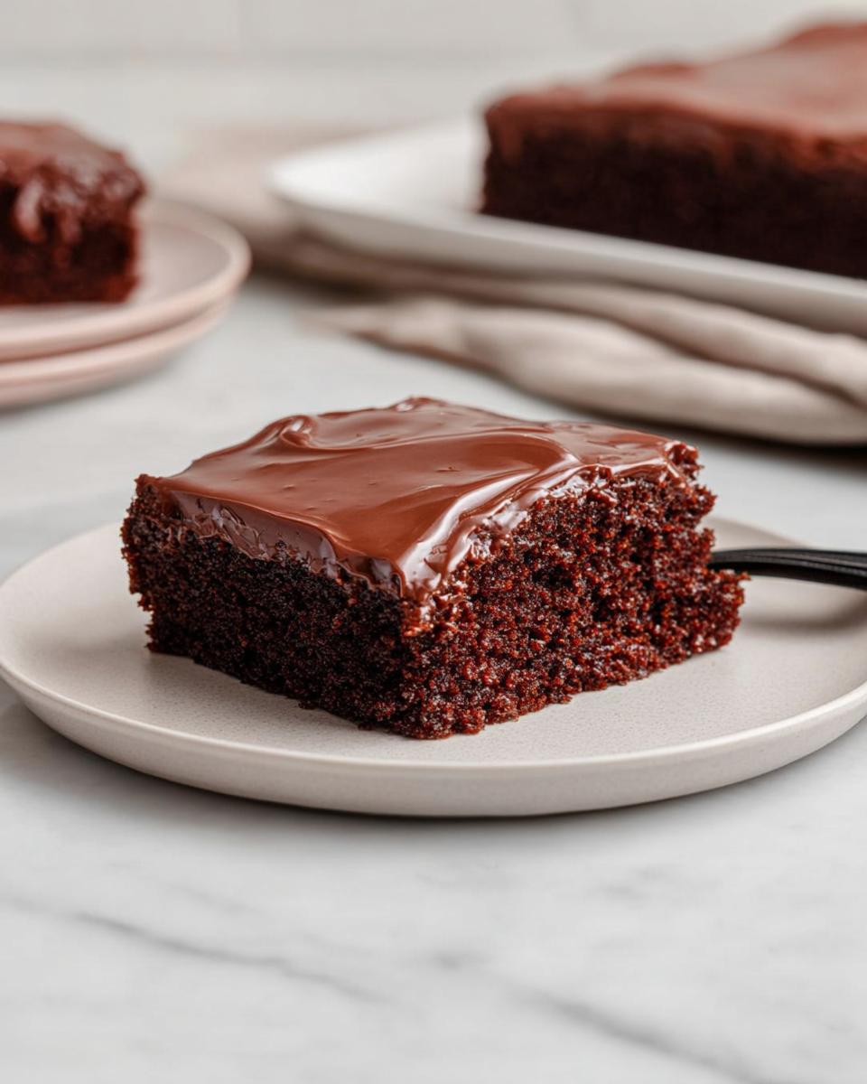 A close-up of a moist slice of Easy Snacking Sheet Cake topped with glossy chocolate frosting on a light plate.