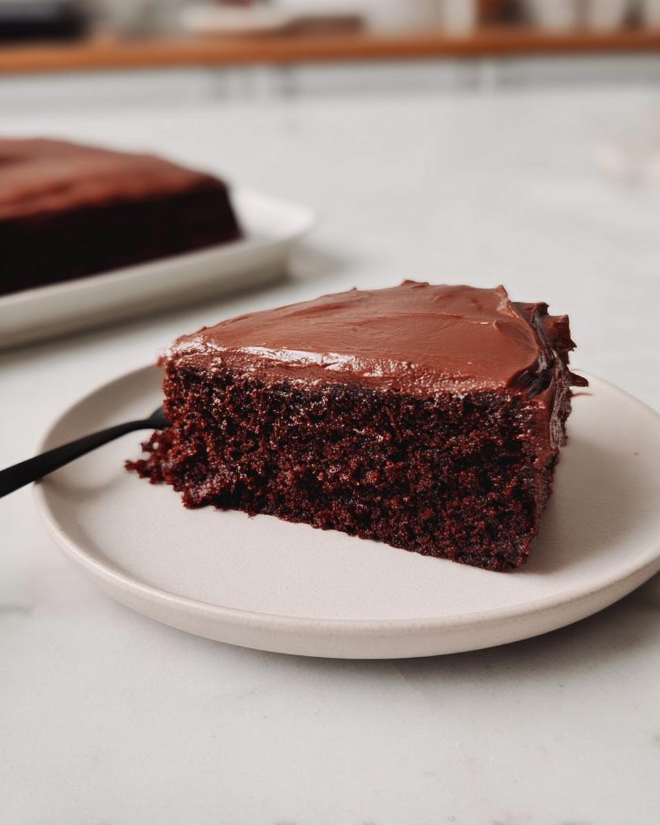 A moist slice of dark chocolate Easy Snacking Sheet Cake topped with glossy chocolate frosting on a white plate.