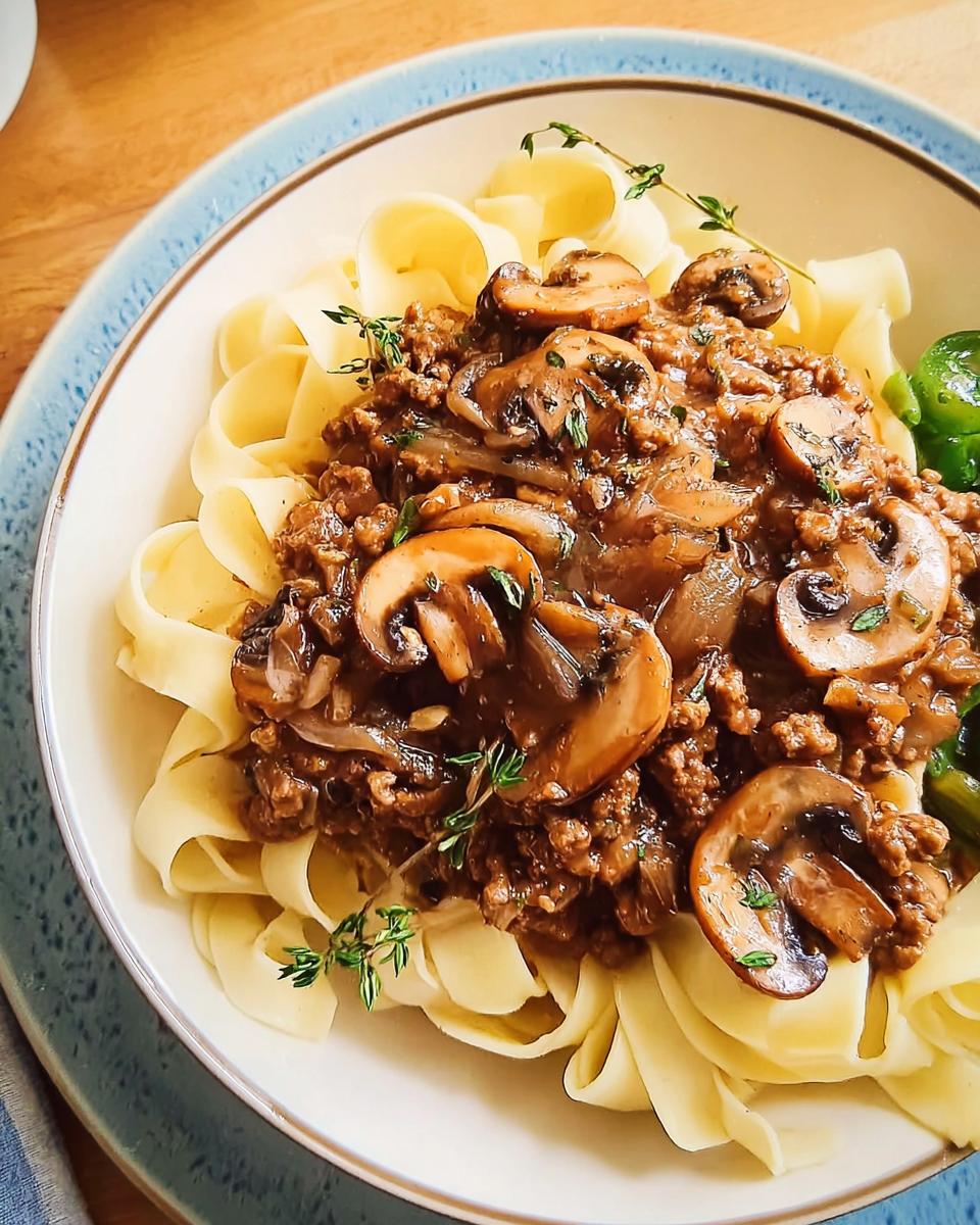 A bowl of wide egg noodles topped with a rich ground beef and mushroom sauce, garnished with fresh thyme. A quick ground beef recipe.