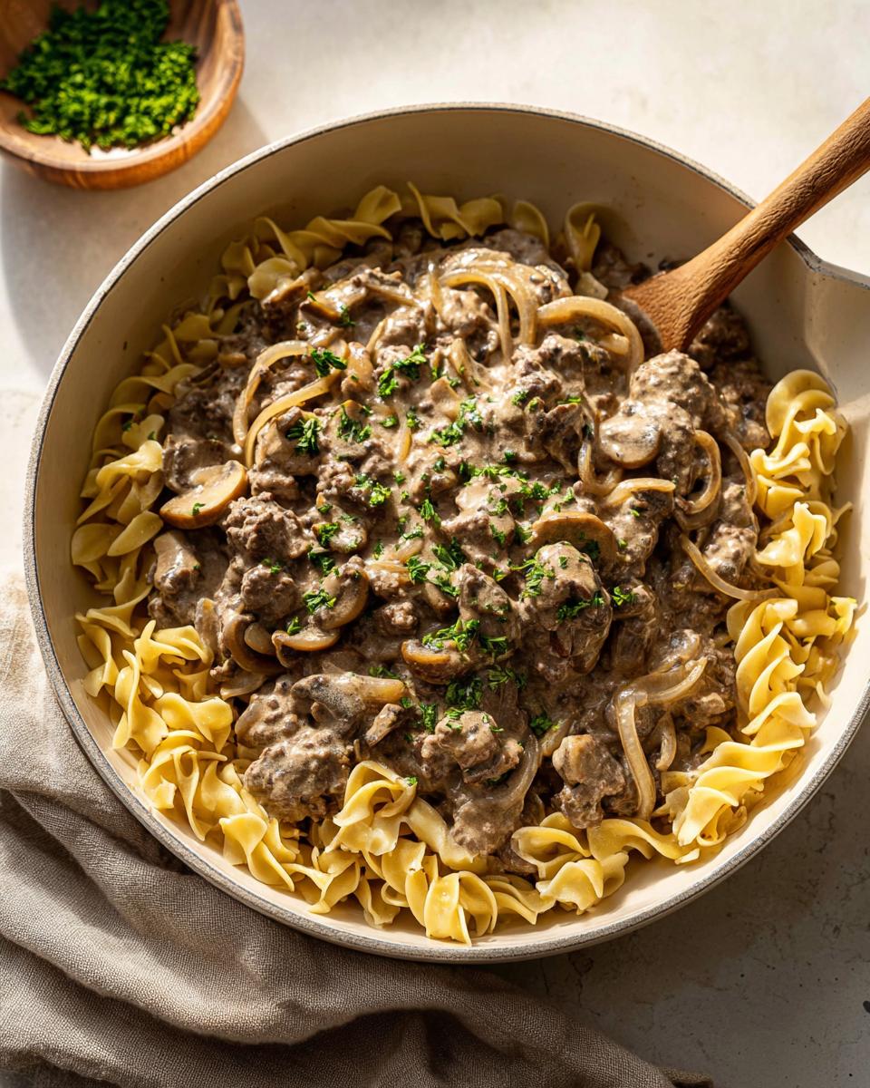 A creamy Ground Beef Stroganoff mixture with mushrooms and onions served over wide egg noodles in a skillet.