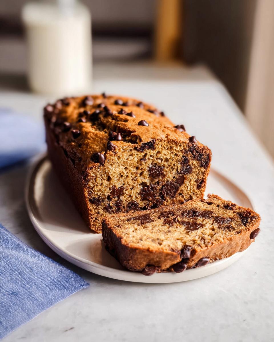 A loaf of moist Healthy Banana Bread (Whole Wheat) topped with chocolate chips, with one slice cut and resting beside it.