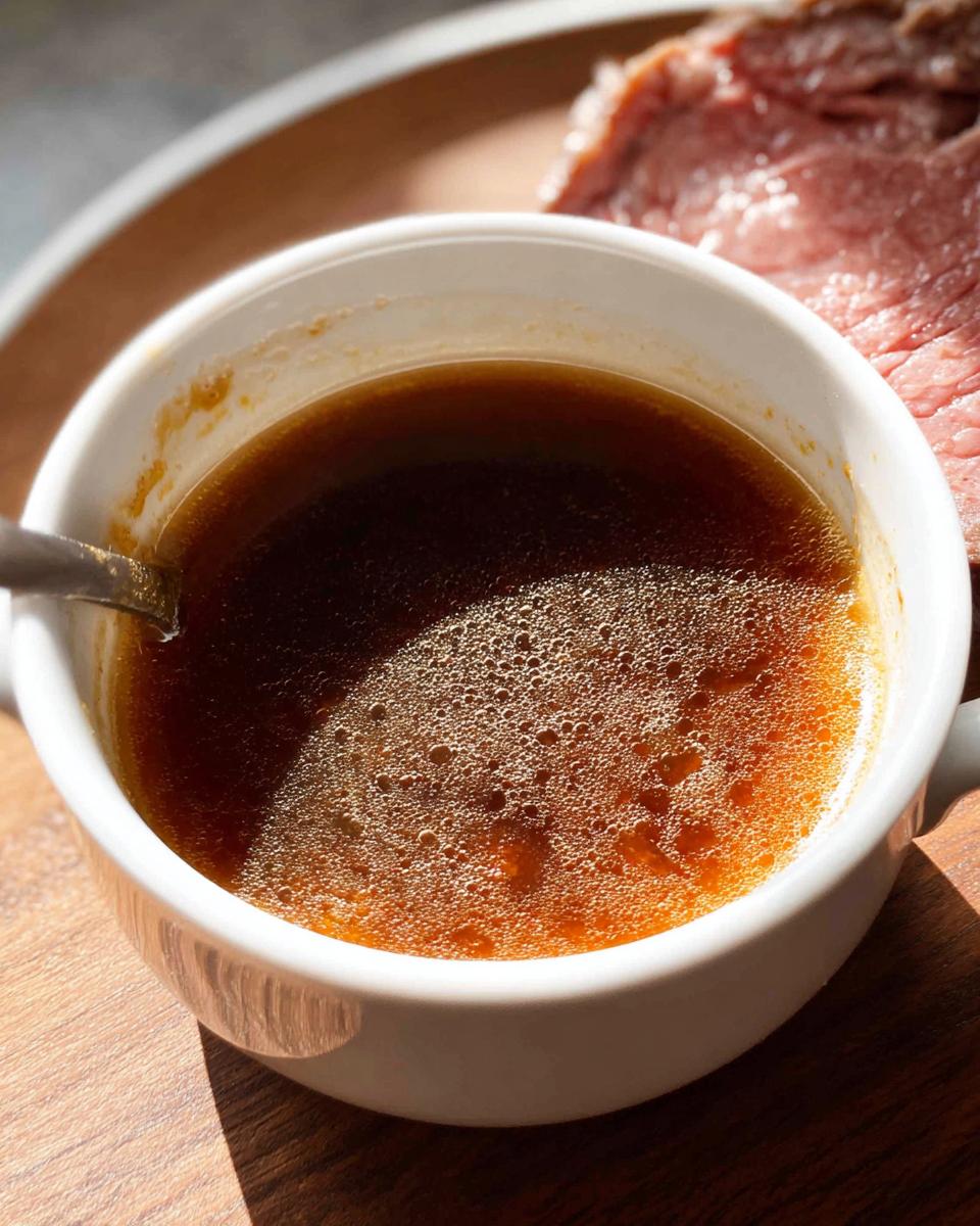 Close-up of rich, brown Homemade Au Jus without drippings served in a white cup next to sliced roast beef.