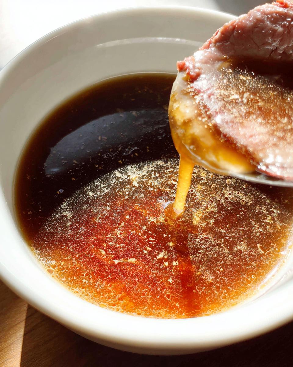 A slice of roast beef being dipped into a bowl of rich, brown Homemade Au Jus without drippings.