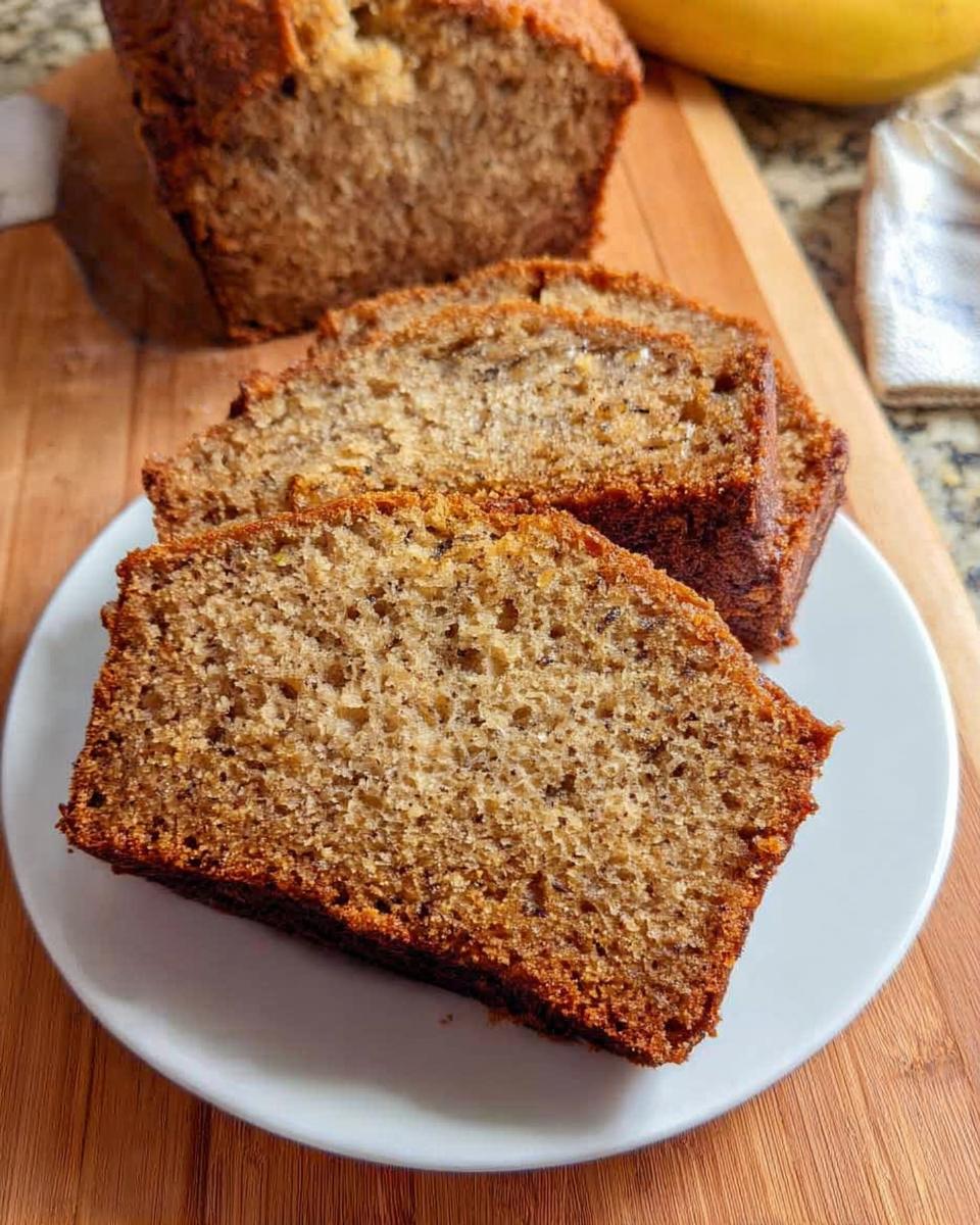 Close-up of three moist slices of One Bowl Banana Bread served on a white plate, with the rest of the loaf in the background.