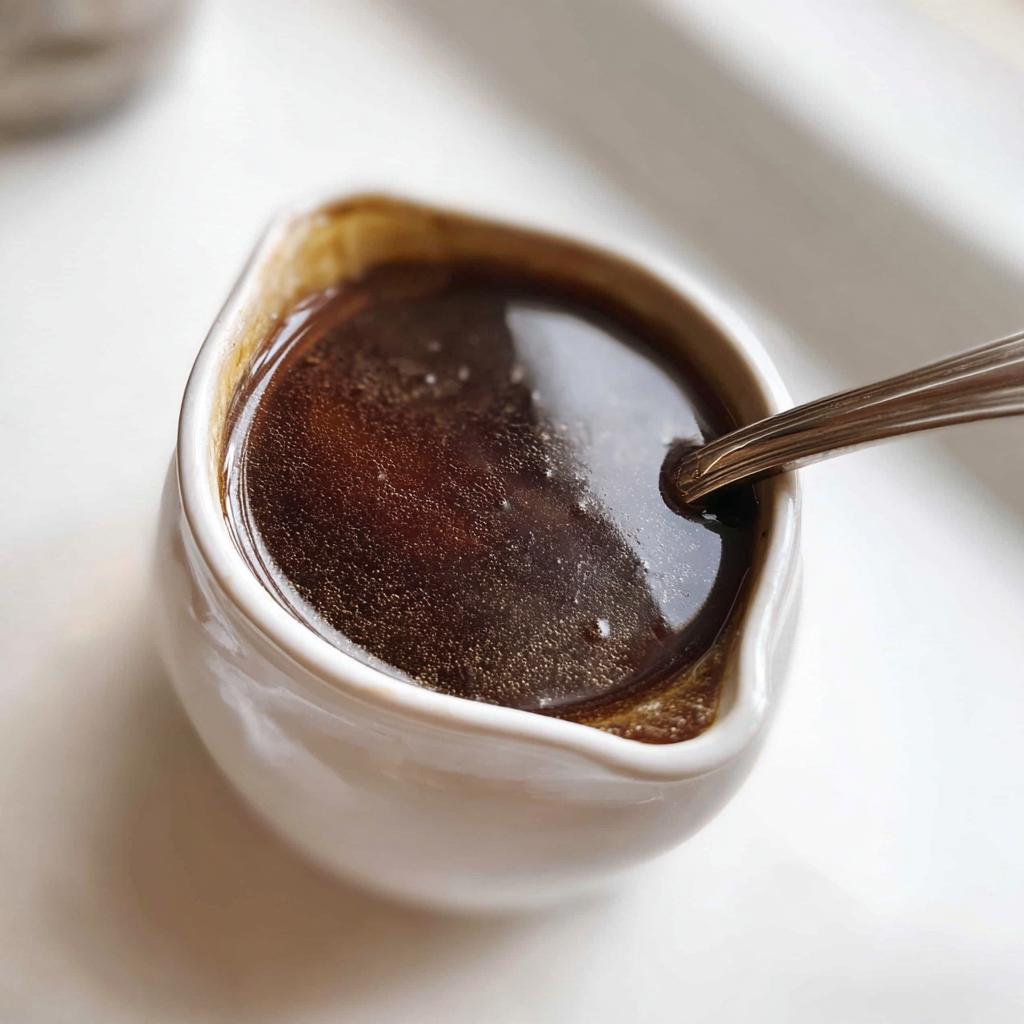 Close-up of rich, dark brown Au Jus Recipe liquid in a small white ceramic gravy boat with a spoon resting inside.