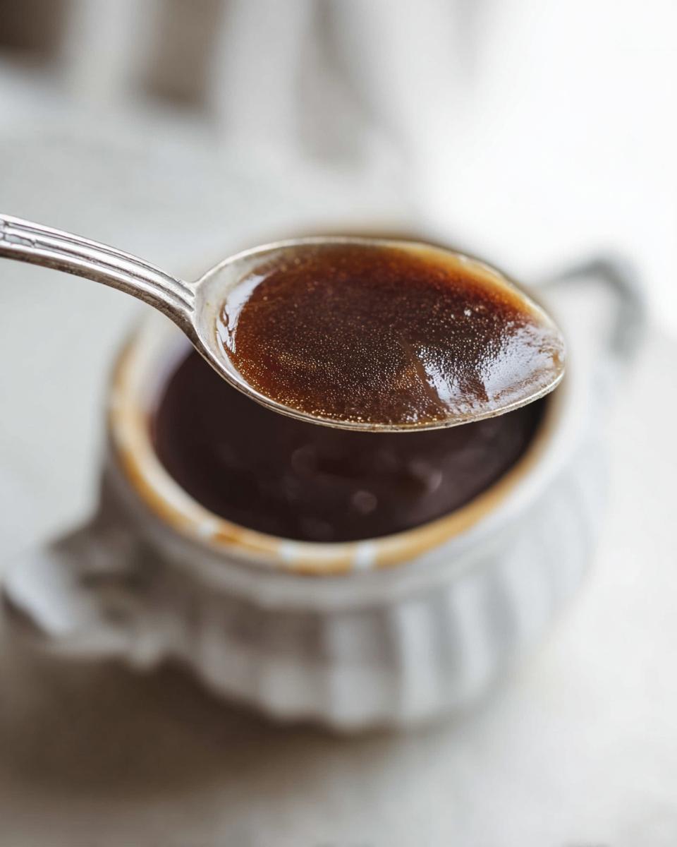 A silver spoon holds a rich, dark brown spoonful of Au Jus recipe, lifted above a small white gravy boat.