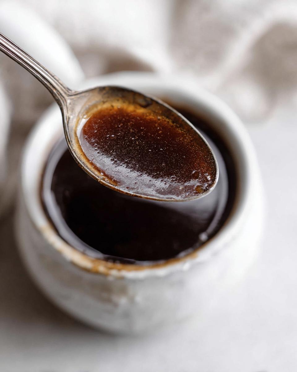 A spoonful of dark, rich Au Jus recipe liquid being lifted from a small white ramekin.