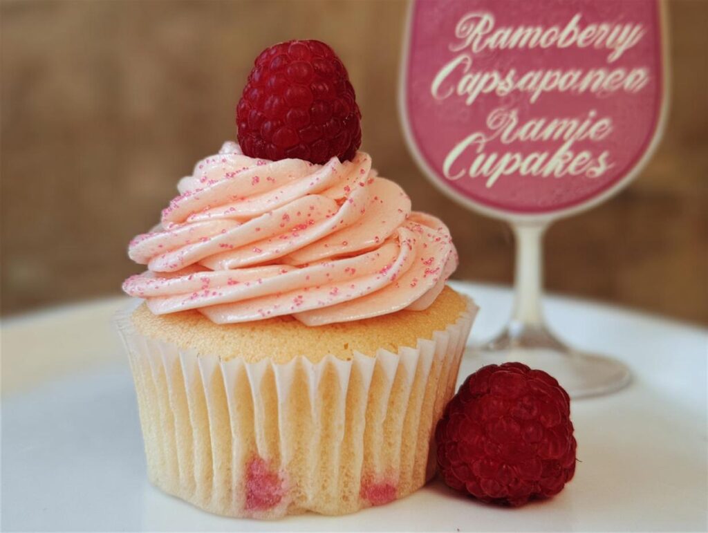 A beautifully frosted Raspberry Champagne Cupcakes topped with a fresh raspberry, with another raspberry beside it.