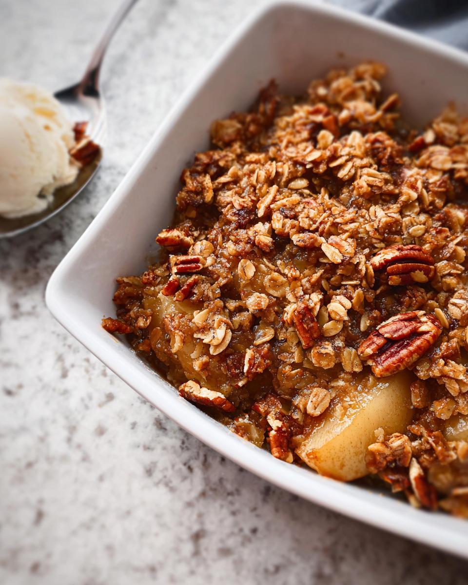 A close-up of a warm pear crisp with a crunchy oat and pecan topping, served with a scoop of vanilla ice cream.