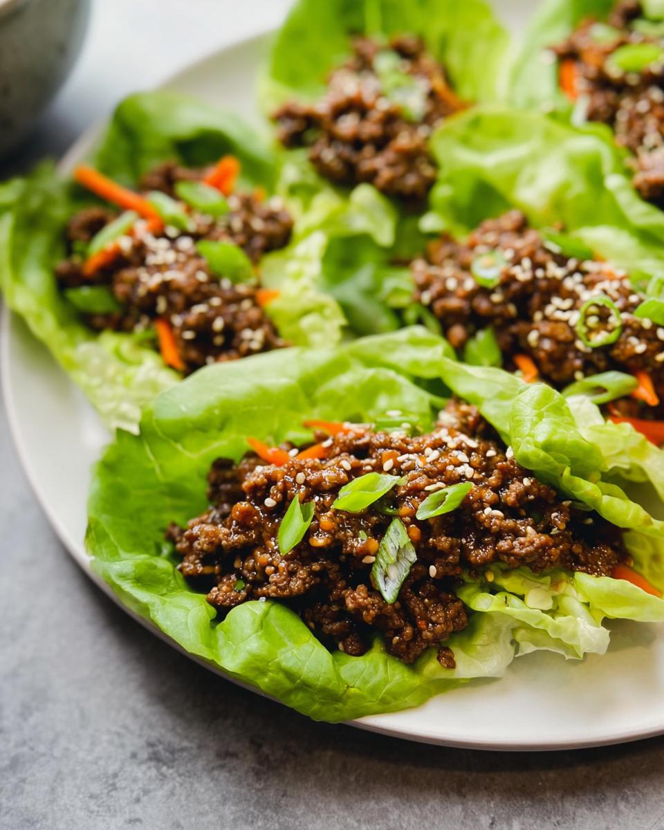 Close-up of several Asian Ground Beef Lettuce Wraps served on a white plate, topped with sesame seeds and green onions.