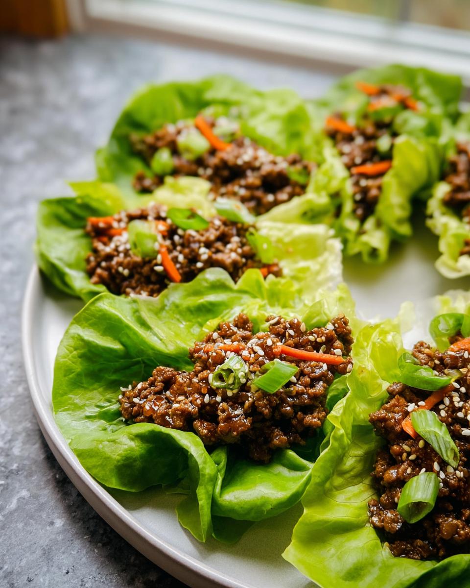 Several Asian Ground Beef Lettuce Wraps served on a light gray plate, topped with sesame seeds and green onions.