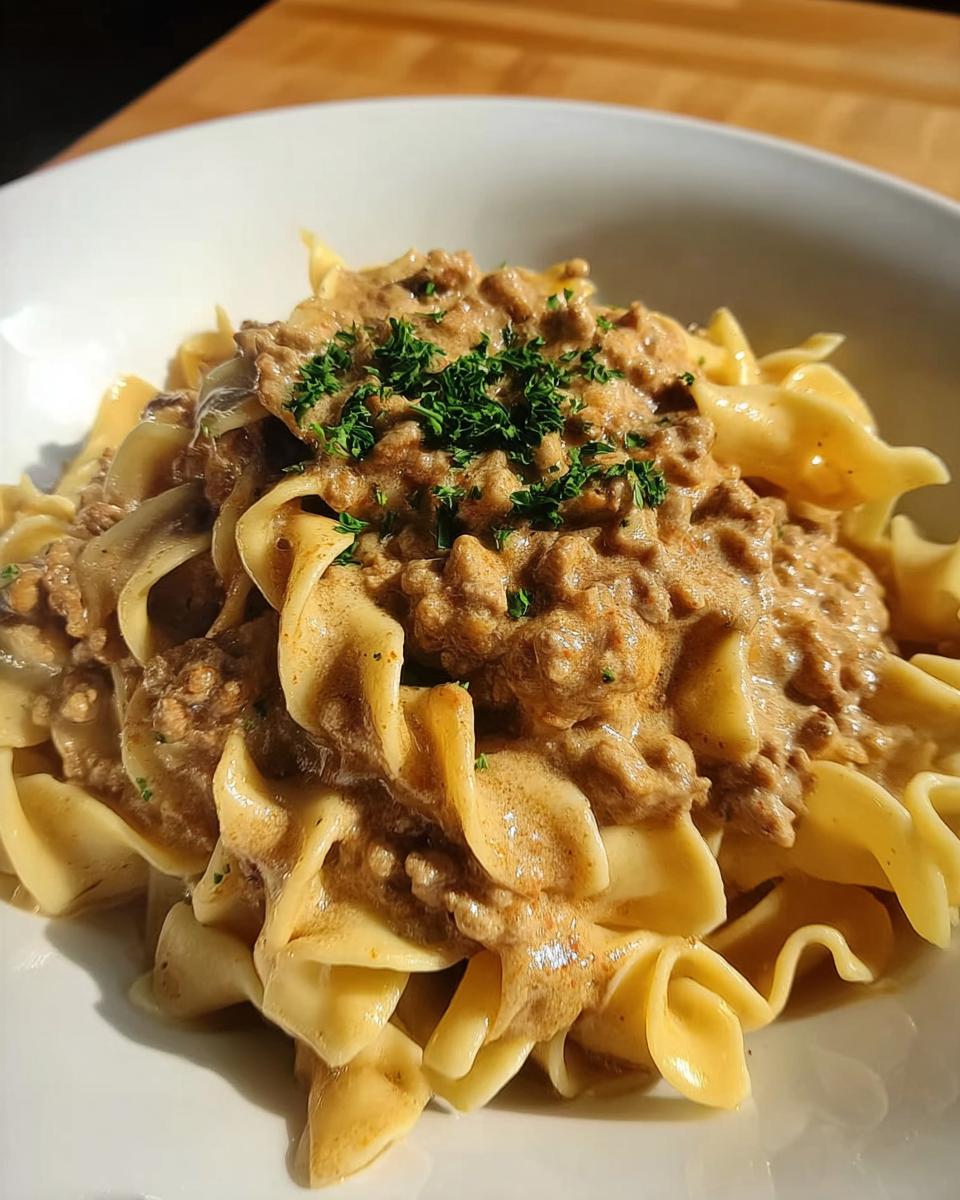 A close-up of a white bowl filled with Creamy Turkey Stroganoff served over wide egg noodles and garnished with parsley.