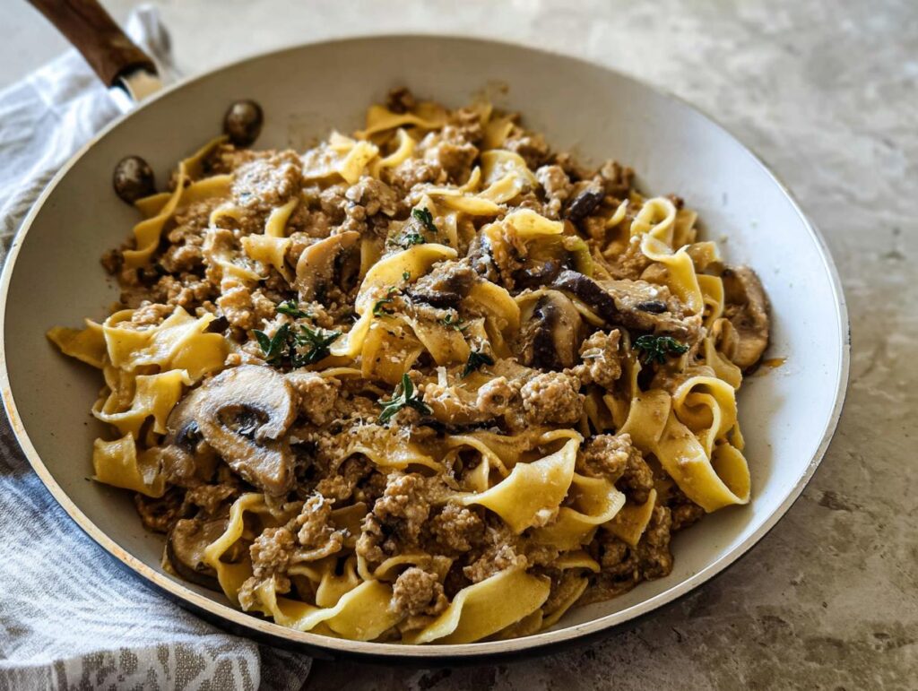 Close-up of Creamy Turkey Stroganoff mixed with wide egg noodles and sliced mushrooms in a light-colored skillet.