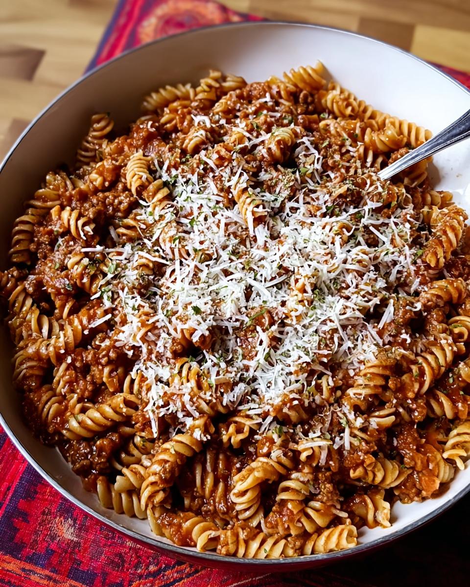A large white bowl filled with rotini pasta tossed in a rich meat sauce, topped generously with grated Parmesan cheese, ready for serving.