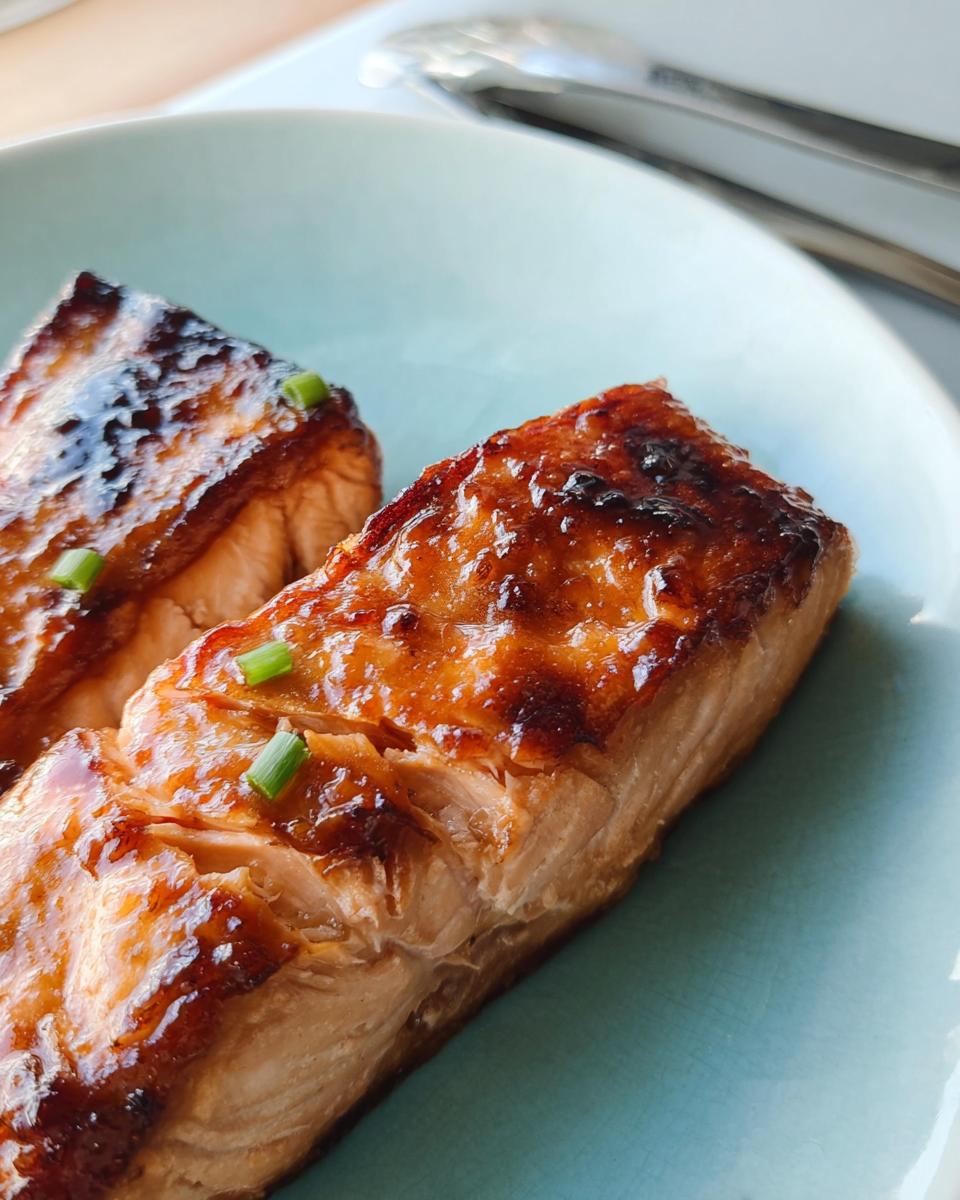 Close-up of two perfectly cooked Miso Glazed Broiled Salmon fillets garnished with green onions on a light blue plate.