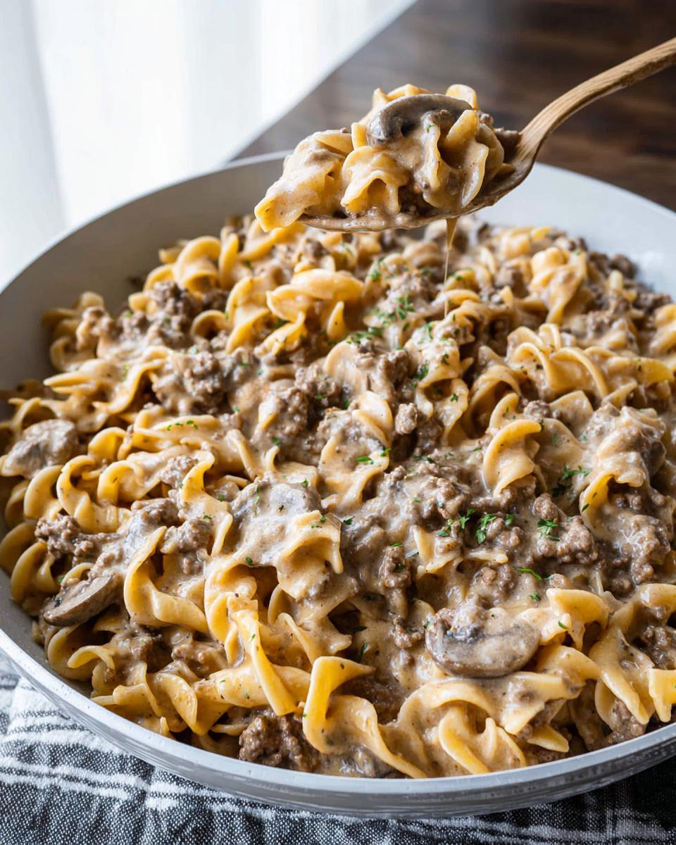 A wooden spoon lifts a portion of creamy One Pot Ground Beef Stroganoff, rich with egg noodles, ground beef, and mushrooms.