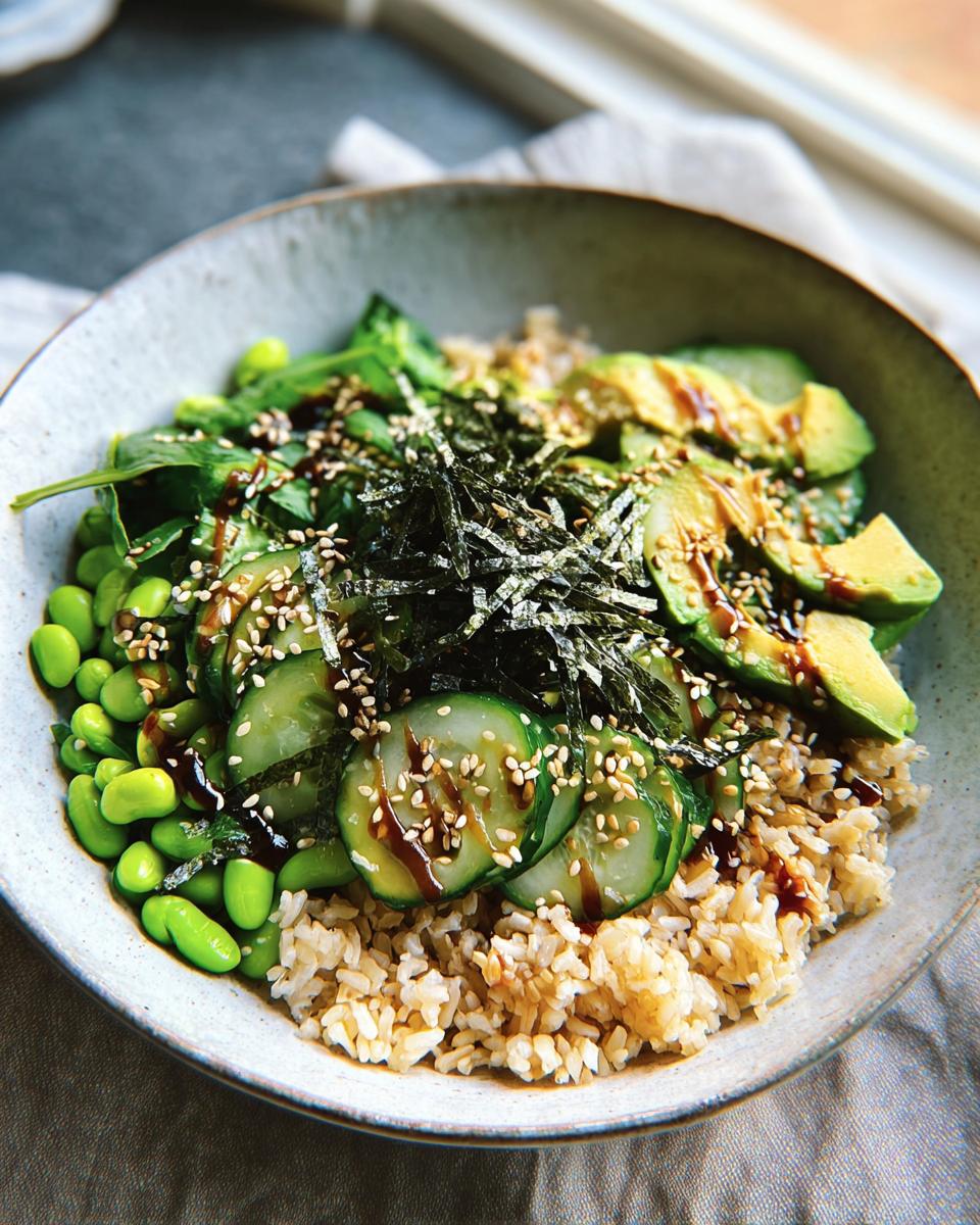 A vibrant rice bowl featuring brown rice, edamame, cucumber slices, avocado, and nori, drizzled with sauce and sprinkled with sesame seeds.