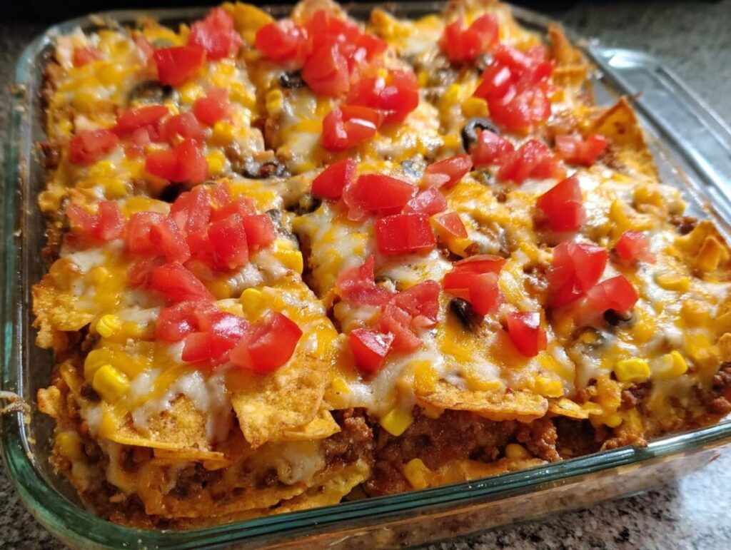 A close-up of a baked Taco Tuesday recipes casserole in a glass dish, topped with melted cheese, corn, and diced tomatoes.