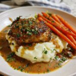 A plate of creamy herb chicken served with mashed potatoes and glazed carrots, drizzled in sauce.