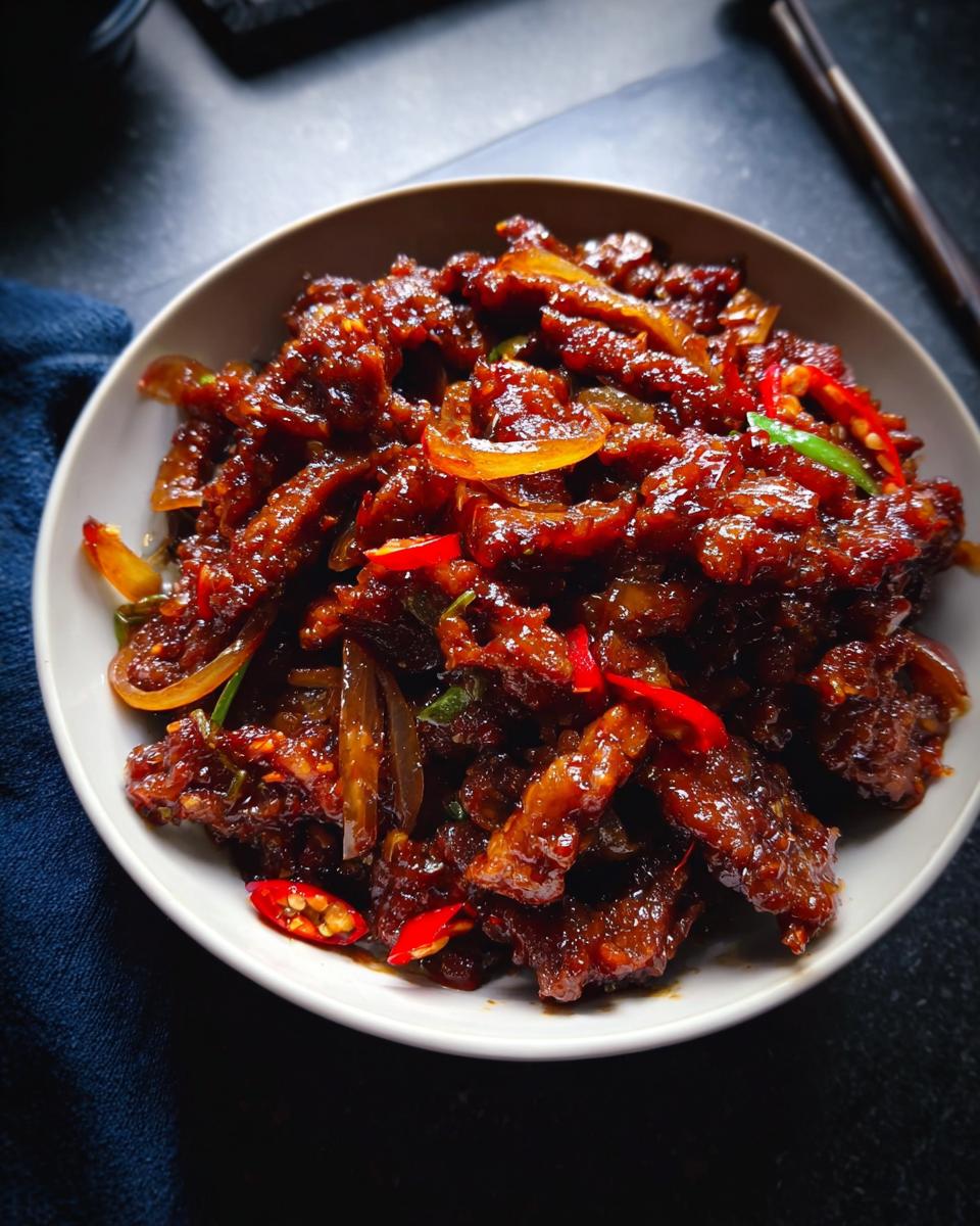 A close-up of a bowl filled with glistening Crispy Chilli Beef Rice, garnished with red chillies and onions.