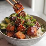 A bowl of white rice topped with glazed crispy salmon pieces, sliced cucumbers, and garnished with sesame seeds and green onions.