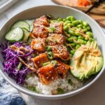 A vibrant Crispy Salmon and Rice Bowl featuring glazed salmon pieces, white rice, edamame, avocado slices, cucumber, and red cabbage, drizzled with sauce.