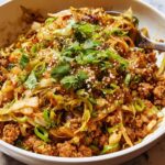 A close-up of a white bowl filled with Egg Roll in a Bowl, featuring ground meat, shredded cabbage, and garnished with green onions and sesame seeds.