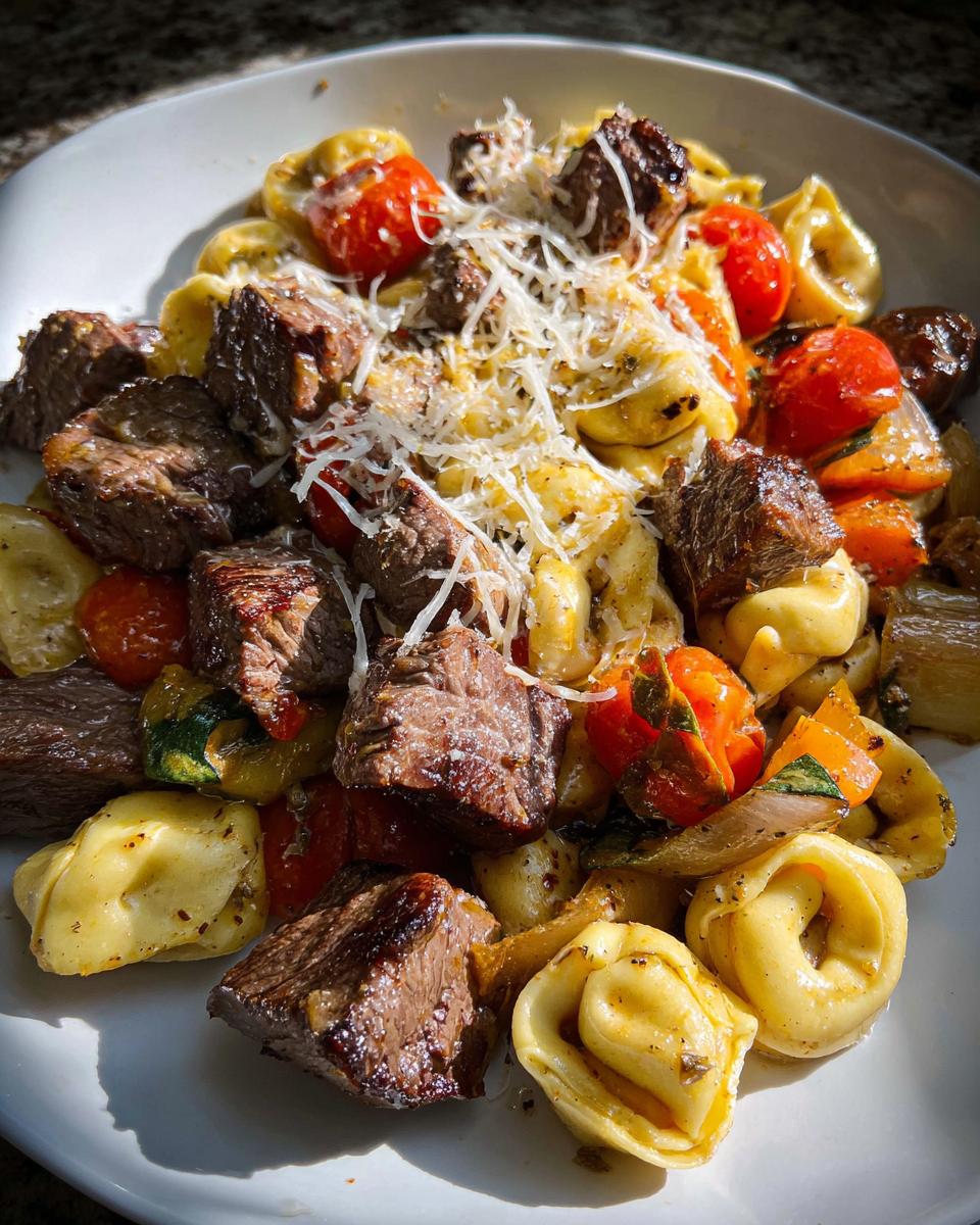 A close-up of a white plate filled with Garlic Steak Tortellini, featuring tender steak bites, tortellini pasta, cherry tomatoes, and shredded cheese.