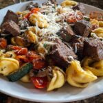 A close-up of a white plate filled with Garlic Steak Tortellini, featuring tender steak, pasta, cherry tomatoes, zucchini, and grated cheese.