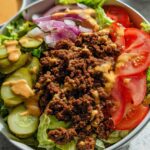 A close-up of a High-Protein Cheeseburger Bowl featuring seasoned ground beef, lettuce, tomato, pickles, red onion, and a creamy sauce.