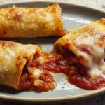 Three HIGH PROTEIN Pepperoni Pizza Rolls on a plate, one is broken open showing melted cheese and pepperoni filling.