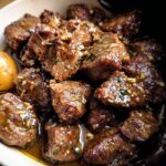 Close-up of tender High Protein Slow Cooker Garlic Butter Beef Bites in a white bowl with a single potato.