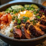 A bowl of Korean BBQ Steak Rice Bowls topped with a sunny-side-up egg, kimchi, cucumbers, and spicy cream sauce.