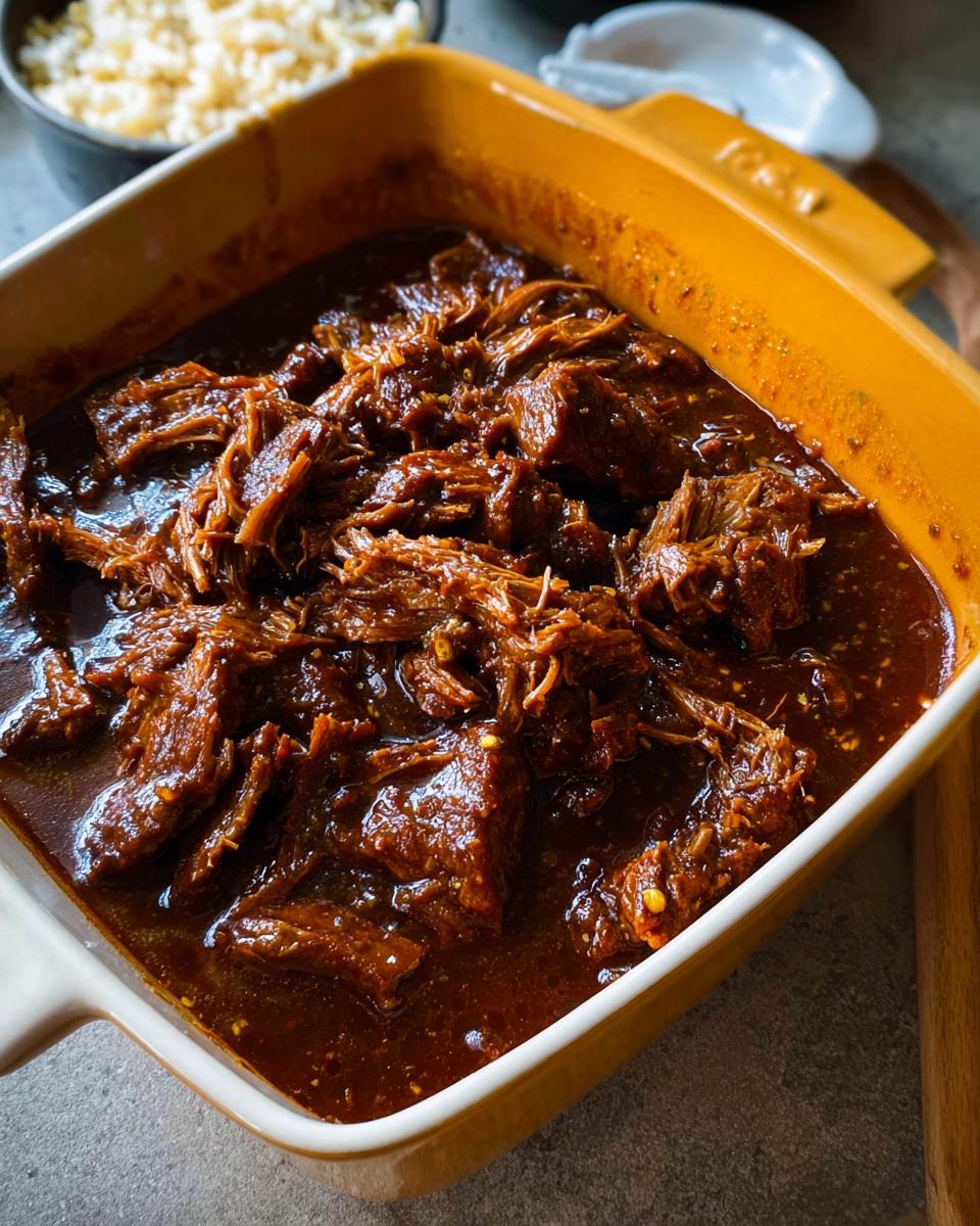 Close-up of shredded Korean Style Pot Roast in a rich, dark sauce within a yellow baking dish.