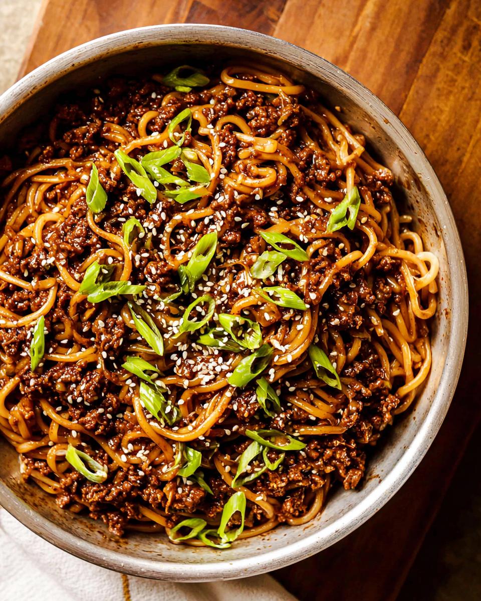 A close-up overhead view of a bowl filled with Mongolian Ground Beef Noodles, garnished with green onions and sesame seeds.