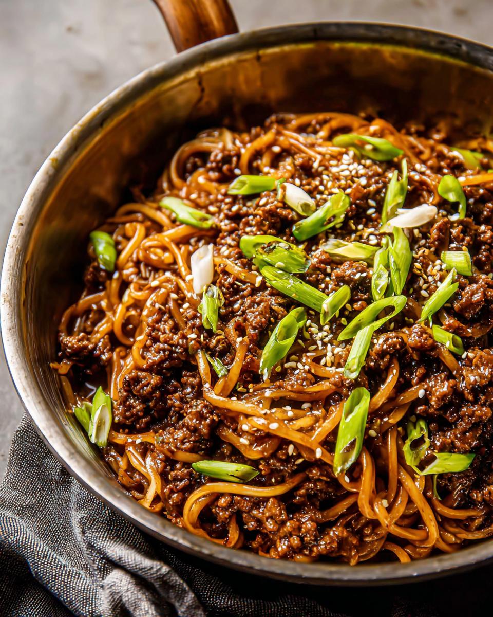 A close-up of a skillet filled with Mongolian Ground Beef Noodles, garnished with green onions and sesame seeds.