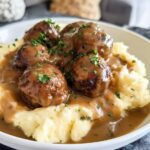 A bowl of Salisbury steak meatballs smothered in gravy, served over garlic herb mashed potatoes and garnished with parsley.
