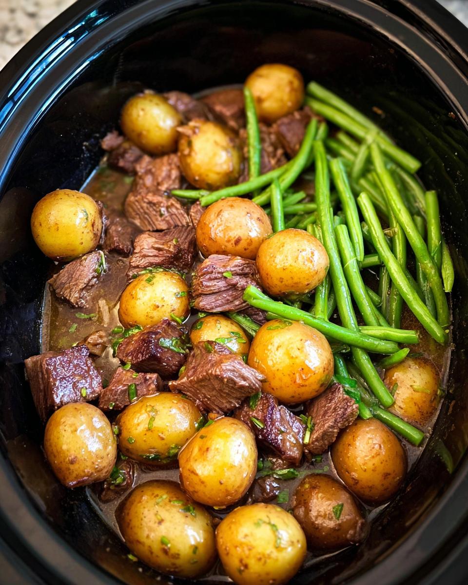 A close-up overhead view of Slow Cooker Garlic Butter Beef with Potatoes and green beans in a black slow cooker.