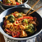 A bowl of Spicy Garlic Chicken and Broccoli Noodle Bowls with thick noodles, chicken, broccoli, and red peppers, topped with sesame seeds.