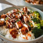 Close-up of a bowl filled with white rice, topped with glazed chicken pieces, roasted broccoli, and a drizzle of creamy sauce.