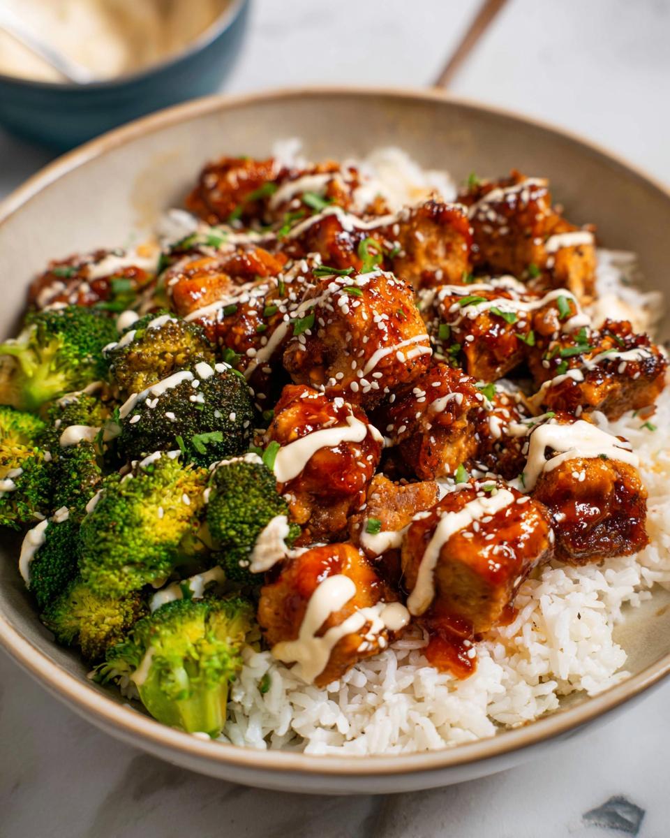 A close-up of a bowl filled with fluffy white rice, topped with glazed sticky chicken pieces and steamed broccoli florets, drizzled with sauce and sesame seeds.