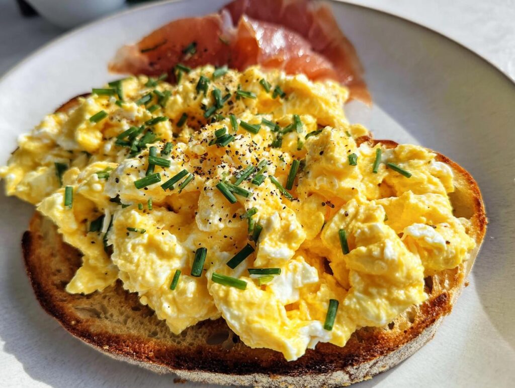 Close-up of Creamy Ricotta Egg Toast topped with fluffy scrambled eggs, chives, and black pepper, served with prosciutto.