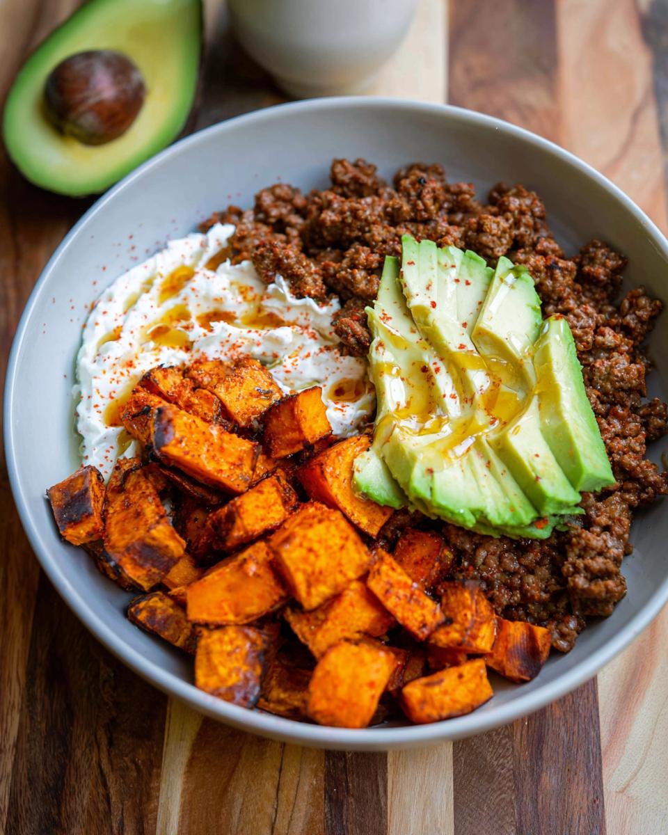 A vibrant Ground Beef Hot Honey Bowl featuring seasoned ground beef, creamy white sauce, roasted sweet potato cubes, and sliced avocado drizzled with honey.