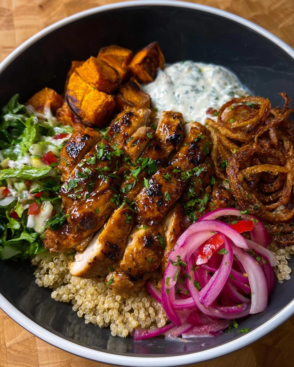 A delicious Hot Honey Chicken Bowl featuring sliced chicken, roasted sweet potatoes, quinoa, salad, pickled onions, and crispy fried onions.