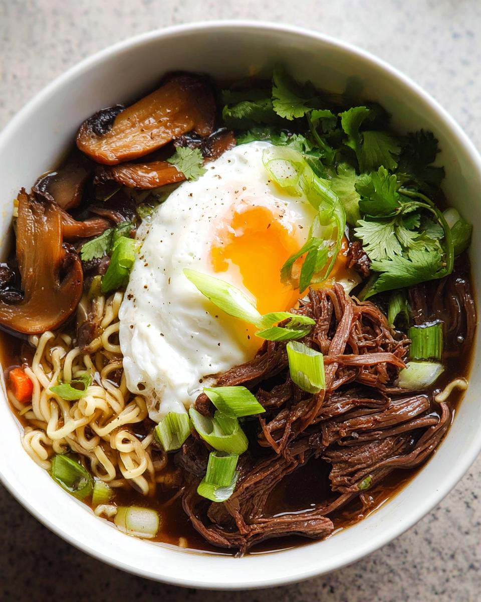 A close-up of a bowl of Slow Cooker Beef Ramen Noodles topped with a fried egg, mushrooms, and green onions.