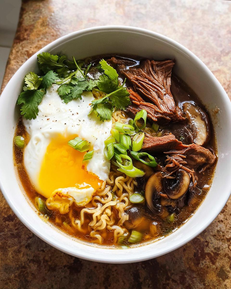 A bowl of Slow Cooker Beef Ramen Noodles topped with a perfectly poached egg, shredded beef, mushrooms, and green onions.